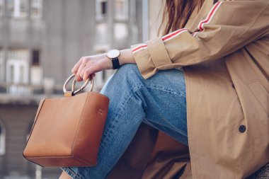 sokak tarzı bej bir palto, denim jeans, ayak bileği botları, kedi göz güneş gözlüğü ve Metal kolu Deri tote çanta çekici bir kadın portresi. moda kıyafet güneşli bir bahar için mükemmel