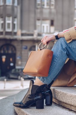 sokak tarzı bej bir palto, denim jeans, ayak bileği botları, kedi göz güneş gözlüğü ve Metal kolu Deri tote çanta çekici bir kadın portresi. moda kıyafet güneşli bir bahar için mükemmel