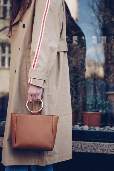 street style portrait of an attractive woman wearing a beige trench coat, denim jeans, ankle boots, cat eye sunglasses and a metallic handle brown tote bag. fashion outfit perfect for sunny spring day