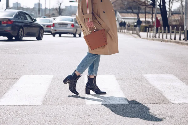 street style portrait of an attractive woman wearing a beige trench coat, denim jeans, ankle boots and metallic handle brown tote bag, crossing the street. fashion outfit perfect for sunny spring day