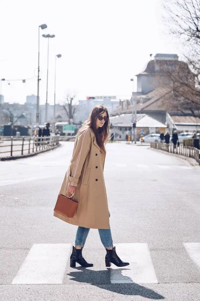 street style portrait of an attractive woman wearing a beige trench coat, denim jeans, ankle boots and metallic handle brown tote bag, crossing the street. fashion outfit perfect for sunny spring day