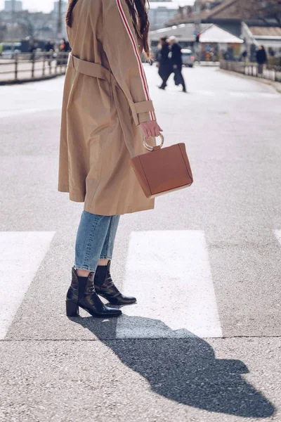 street style portrait of an attractive woman wearing a beige trench coat, denim jeans, ankle boots and metallic handle brown tote bag, crossing the street. fashion outfit perfect for sunny spring day