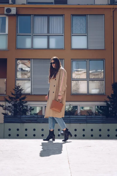 street style portrait of an attractive woman wearing a beige trench coat, denim jeans, ankle boots and metallic handle brown tote bag, crossing the street. fashion outfit perfect for sunny spring day