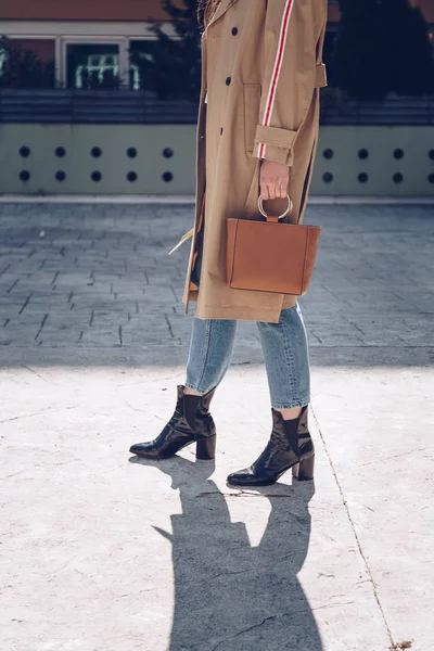 street style portrait of an attractive woman wearing a beige trench coat, denim jeans, ankle boots and metallic handle brown tote bag, crossing the street. fashion outfit perfect for sunny spring day