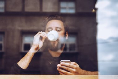 Cep telefonunuzun yazarak bir kahve bar, kahve içme otantik ve çekici bir adam portresi. Şehir yansımaları ile pencere camı vurdu. Cep telefonunu kullanarak serbest meslek. Sosyal ağ, web tarama, manifatura.