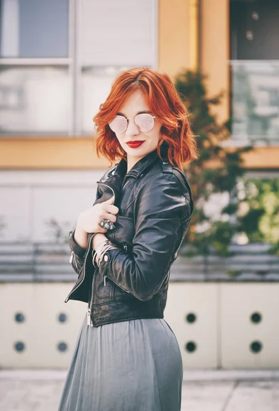 street style fashion portrait, fashionable ginger hair young woman posing in  the city wearing leather jacket and round sunglasses