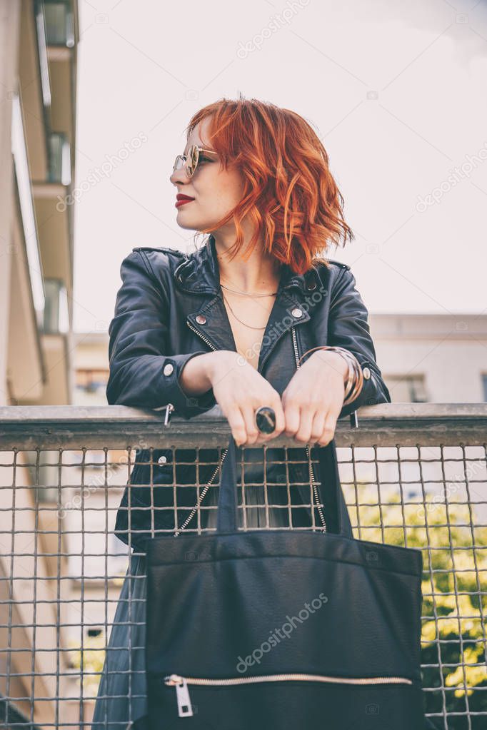 street style fashion potrait of a  beautiful young woman wearing a leather jacket, round sunglasses and holding a black handbag