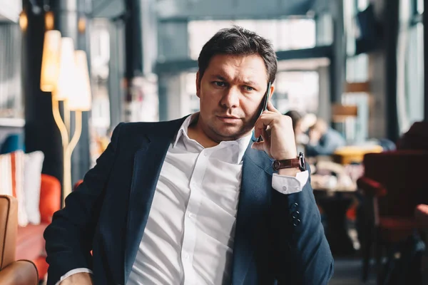 Young good looking male manager sitting in a restaurant while having a business call with his partner, holding a smartphone. Concept of working on a break.