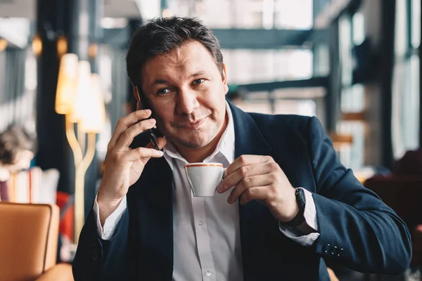 Genç ve kahve içme ve bir iş olması gülümseyen bir restoranda oturan iyi görünümlü erkek bankacı onun cep telefonu çağrı.