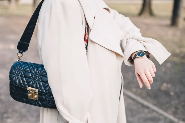 Closeup of young woman in white coat, looking at time on her wrist watch and wearing black quilted handbag.