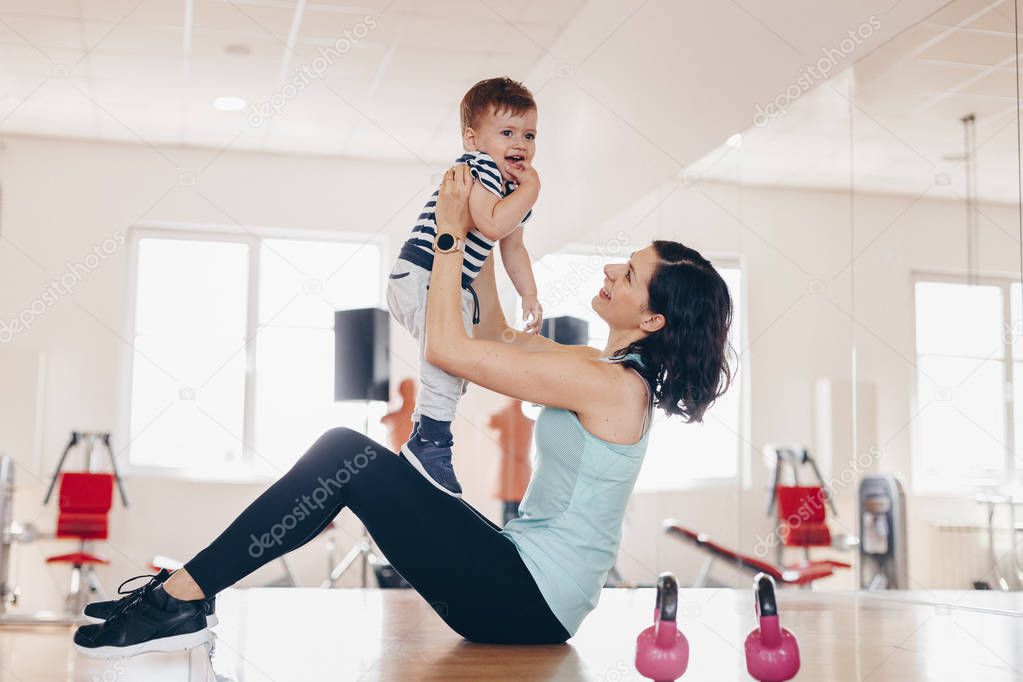 Portrait of happy  young fit mother doing exercise at gym and having fun with her baby boy. Concept of healthy living.  Sport, motherhood and active concept.