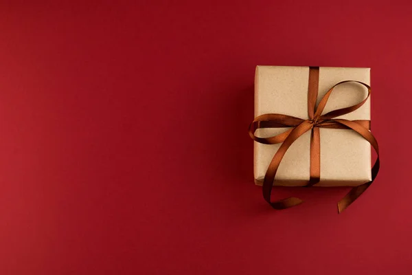 A gift square box packed in craft paper and tied with a brown ribbon on a red background close-up. Copy space.