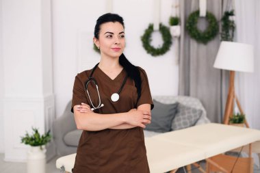 Kameraya bakarak ve modern tıbbi ofis masaj masa arkasında, stetoskop ile gülümseyen genç kadın doktor. Sağlık ve tıp kavramı.