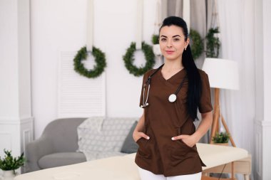 Kameraya bakarak ve modern tıbbi ofis masaj masa arkasında, stetoskop ile gülümseyen genç kadın doktor. Sağlık ve tıp kavramı.