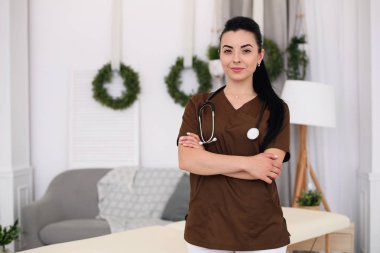 Kameraya bakarak ve modern tıbbi ofis masaj masa arkasında, stetoskop ile gülümseyen genç kadın doktor. Sağlık ve tıp kavramı.