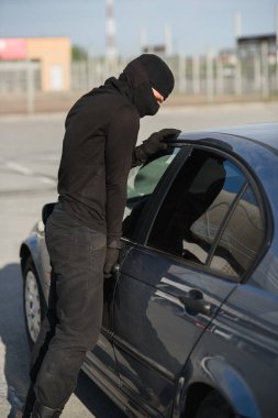 An offender in black clothes and mask is trying to steal a car in a parking lot. Stop criminality. clipart