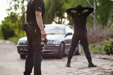 Polis memuru kelepçe elinde tutar. Maskeli tutuklanan suçlu üzerinde kafa katlanmış elleriyle geri döndü.