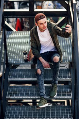 Genç yakışıklı hipster adam endüstriyel bina demir merdivenlerde smartphone kullanarak kaykay ile.