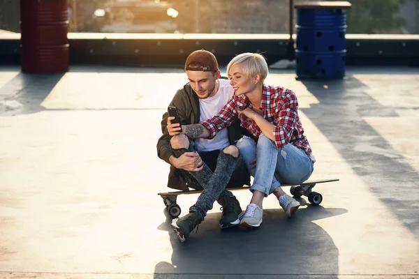 英俊时尚的情侣坐在长板滑板上的屋顶游乐场的工业大厦和使用智能手机 现代城市活跃生活方式概念 — 图库照片