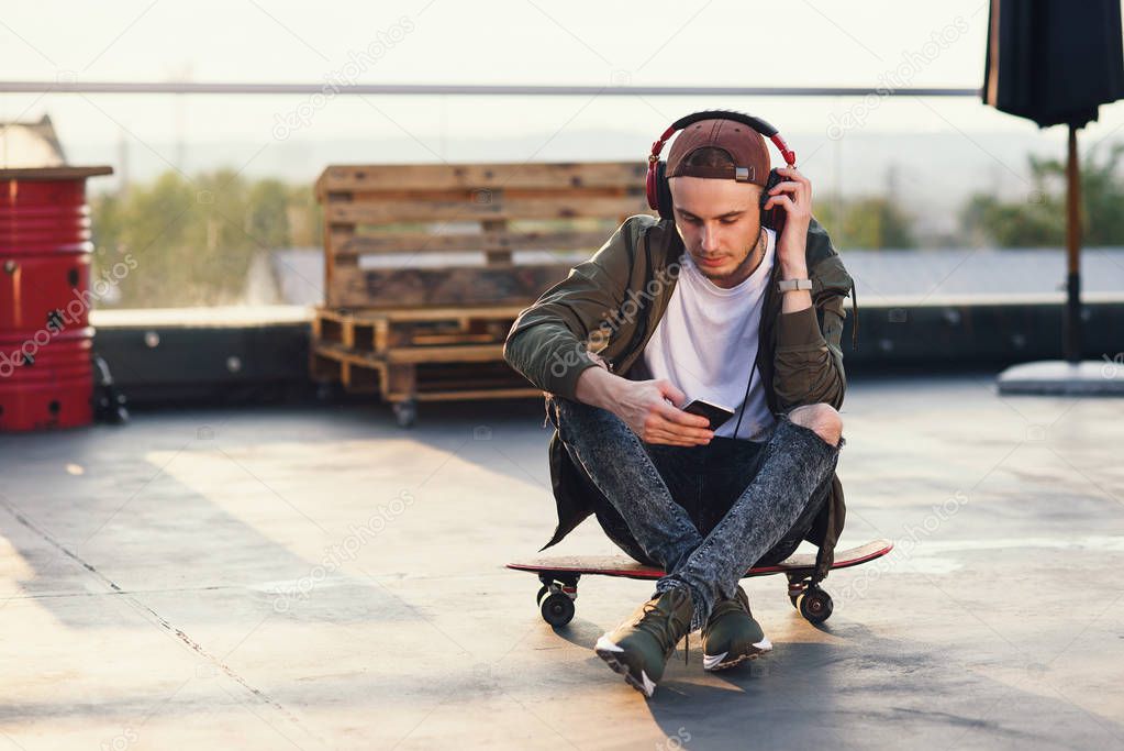 Young handsome man listening music in headphones and using smartphone while sitting on skateboard on the rooftop of industrial building at the sunset.