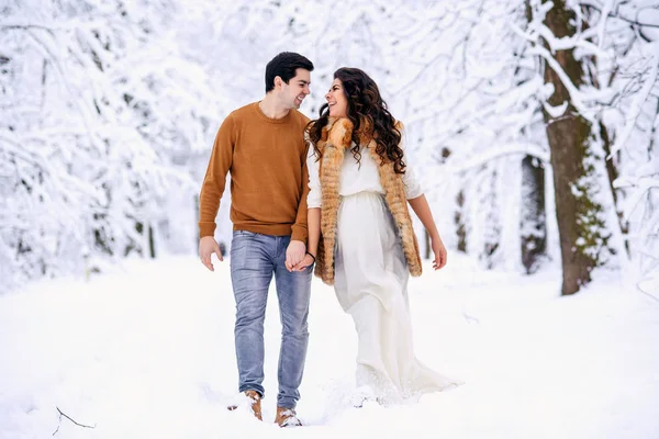 美丽的孕妇穿着一件长裙 穿着一件轻薄的外套 穿着毛衣和牛仔裤走在雪堆的冬季公园里 家庭的爱和关怀概念 — 图库照片