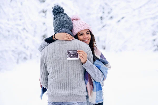Beautiful pregnant woman in warm pink hat hugs gently her husband and holds ultrasonic pregnant picture in her hand. Family love and care concept.