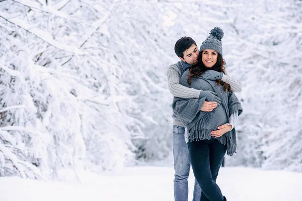 丈夫穿着灰色毛衣和牛仔裤 温柔地抱着美丽怀孕的妻子 穿着灰色针织毛衣 围巾和牛仔裤在雪冬公园 怀孕的概念 — 图库照片