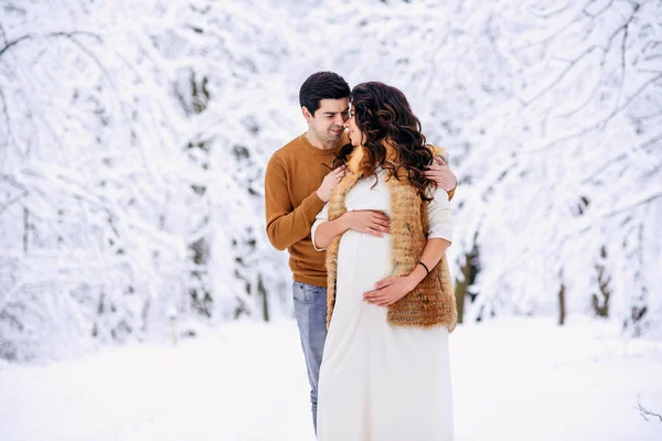 丈夫穿着毛衣和牛仔裤 温柔地抱着他美丽的怀孕妻子 穿着一件长裙 穿着一件外套 穿着雪天的冬日公园 家庭的爱和关怀概念 — 图库照片