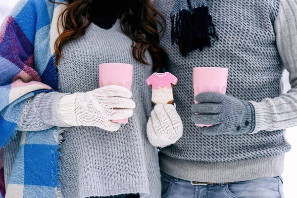 Warm dressed hands of young couple holds pink cups and pink biscuits. Close up.