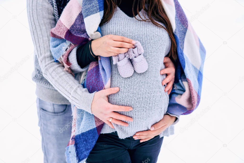 Close up man holding woman's pregnant belly and the woman holding baby shoes in her hand while they stand on snowy winter park.