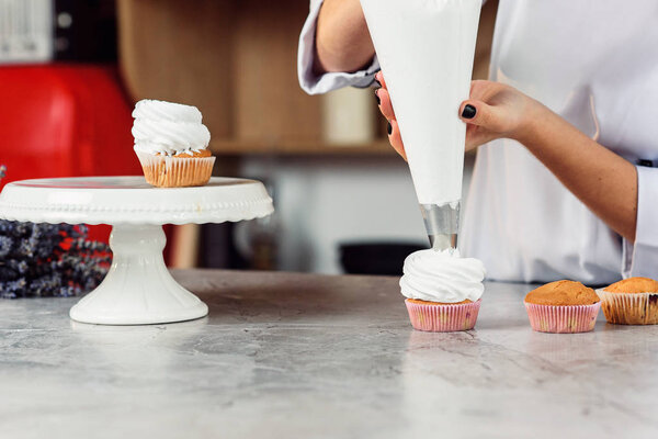Close up hands of the female chef with confectionery bag squeezing cream on cupcakes.