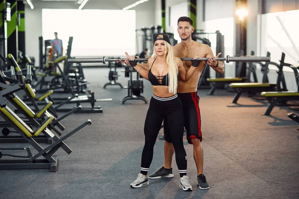 Beautiful girl doing squats under the supervision of the coach. Young couple is working out at gym. Attractive woman and handsome muscular man training together in modern gym.
