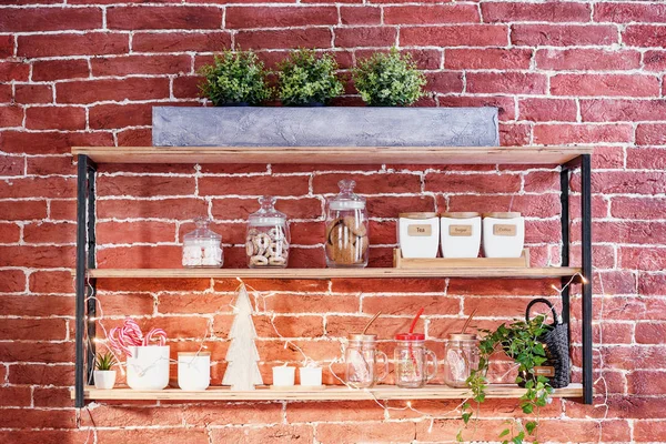 Mutfak aksesuarları ile şık loft tarzı ahşap raf. Çay, kahve ve şeker, şeffaf cam bardak, tatlılar ve dekoratif ahşap Noel ağacı tuğla duvardaki raflarda saklamak için seramik kavanoz.