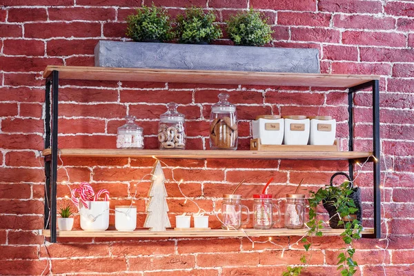Mutfak aksesuarları ile şık loft tarzı ahşap raf. Çay, kahve ve şeker, şeffaf cam bardak, tatlılar ve dekoratif ahşap Noel ağacı tuğla duvardaki raflarda saklamak için seramik kavanoz.
