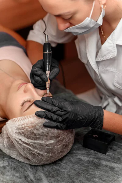 Permanent brow makeup in beauty salon, closeup. Professional beautician doing eyebrow tattoo at woman face.  Cosmetology.