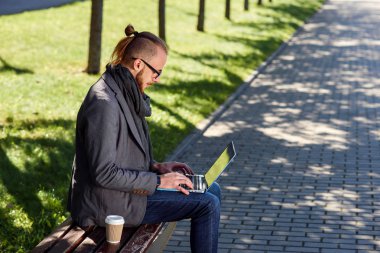 Beyaz genç adam tezgah laptop ve kahve ile oturmuş sakallı kapatın. Parkta bankta oturmuş ve dizüstü bilgisayar kullanarak öğrenci.