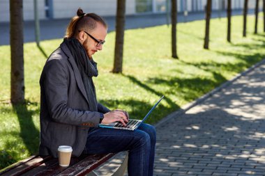 Ahşap Bank laptop ve kahve ile oturmuş sakallı yakışıklı beyaz iş adamı. Parkta bankta oturmuş ve metin dizüstü bilgisayarda yazarak genç erkek öğrenci.
