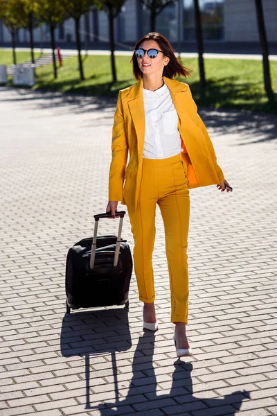 Attractive business woman in stylish yellow suit pulls a suitcase, hurries to a business meeting. Attractive business woman going on a business trip pulling her suitcase along the sidewalk behind her.