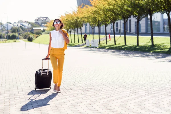 Attractive business woman in stylish yellow suit pulls a suitcase, hurries to the airport. Attractive business woman going on a business trip pulling her suitcase behind her.