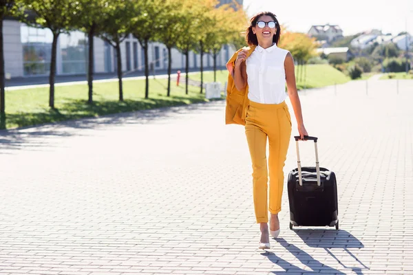 Attractive business woman in stylish yellow suit pulls a suitcase, hurries to the airport. Attractive business woman going on a business trip pulling her suitcase behind her.