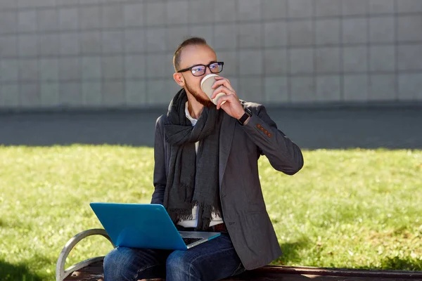 Eşarp içecekler kahve ve açık havada İş Merkezi yakınındaki Park ahşap bankta oturmuş bir öğle yemeği molası dizüstü kullanarak ceket giymiş gözlüklü genç işadamı.