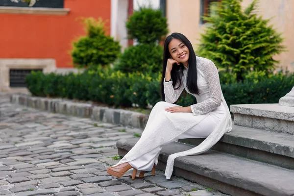 Gros plan portrait de belle fille asiatique habillée en robe blanche Ao Dai assis sur les escaliers dans la cour . — Photo