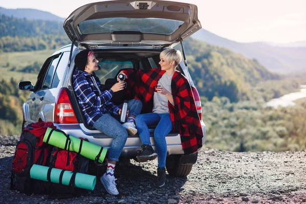 Viajar, turismo e conceito de amizade. Jovens amigos viajantes sentados no porta-malas do carro e bebem chá quente. Duas meninas hipster felizes desfrutando de férias nas montanhas . — Fotografia de Stock