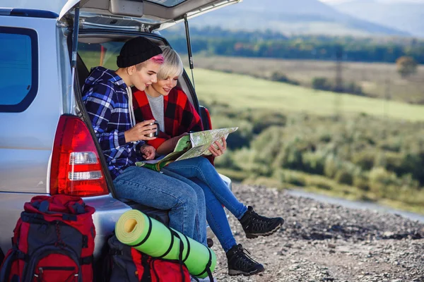 Jovens amigos viajantes sentados no porta-malas do carro com boné de chá e olhando para o mapa de papel. Duas meninas hipster felizes desfrutando de férias nas montanhas. Verão conceito de viagem . — Fotografia de Stock