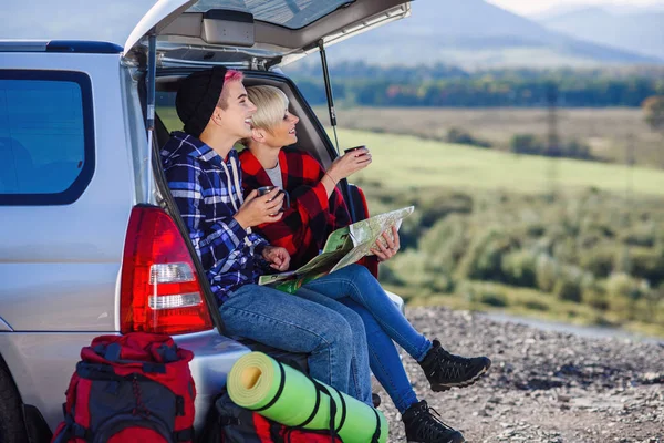 Jovens amigos viajantes sentados no porta-malas do carro com boné de chá e olhando para o mapa de papel. Duas meninas hipster felizes desfrutando de férias nas montanhas. Verão conceito de viagem . — Fotografia de Stock