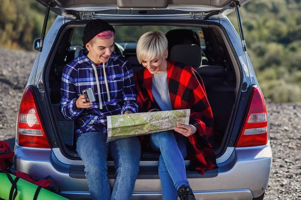 Jovens amigos viajantes sentados no porta-malas do carro com boné de chá e olhando para o mapa de papel. Duas meninas hipster felizes desfrutando de férias nas montanhas. Viagem de verão . — Fotografia de Stock