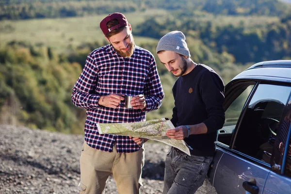 Dois amigos viajantes com boné de chá e olhando para o mapa de papel para encontrar o caminho correto. Homens hipster felizes a explorar a localização nas montanhas. Verão conceito de viagem . — Fotografia de Stock