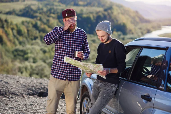 Two friends travelers with cap of tea and looking at the paper map to find correct way. Happy hipster men exploring location in mountains. Summer travel concept.