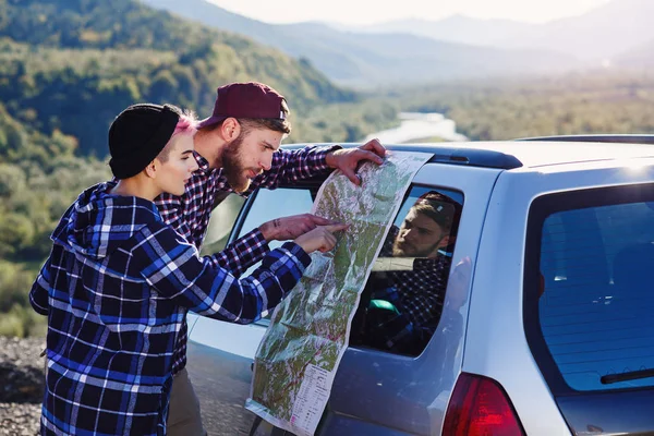 Casal turístico feliz com mapa de papel perto de carro. Jovens sorridentes usando mapa. Viajar de carro alugado em férias de verão. Hipster homem e mulher tendo viagem ao ar livre nas montanhas . — Fotografia de Stock