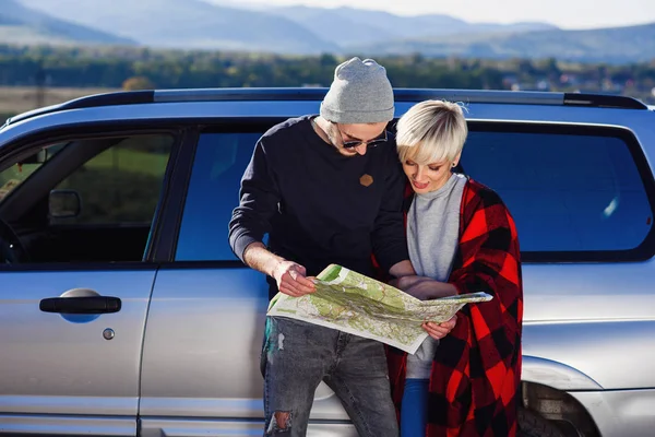 Casal turístico feliz com mapa de papel perto de carro alugado. Jovens na moda usando mapa. Viajar de carro em férias de verão em montanhas. Hipster homem e mulher tendo viagem ao ar livre . — Fotografia de Stock